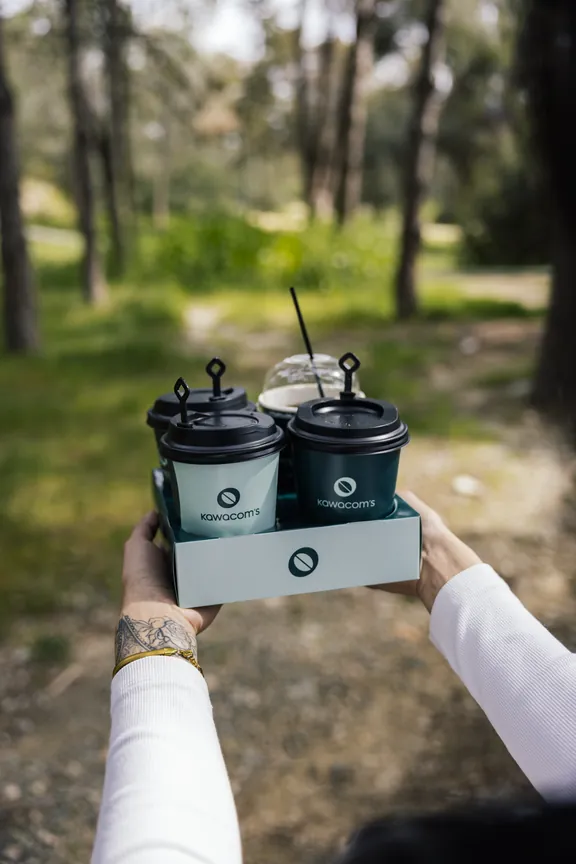 Hand carrying two Kawacom's coffees in a cup holder outdoors